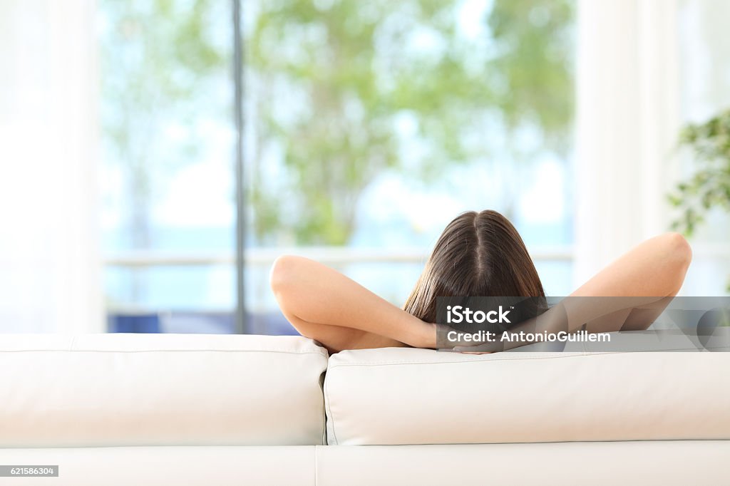 Woman relaxing on a couch at home Rear view of a woman relaxing sitting on a couch with the hands on the head and looking outdoors through the window of the livingroom at home Freshness Stock Photo