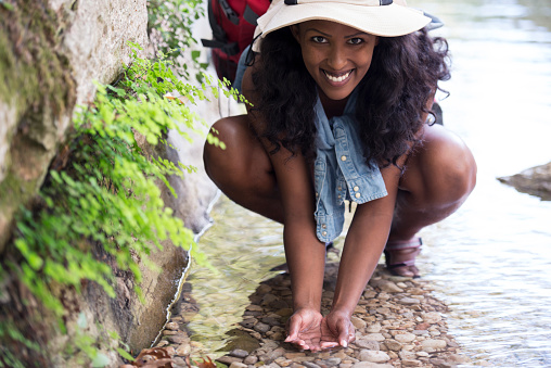 Backpacker tourist woman taking fresh water from river. Woman ready to drink and refresh her face with the spring natural water from a mountain river. Woman wearing short shorts, tank top, fishing hat and red backpack, looking over the camera with a smile.