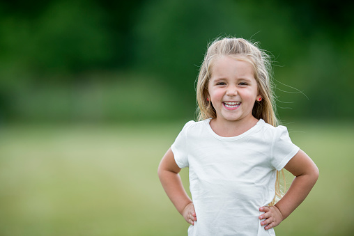 A cute preschool age girl is standing outside at the park and is smiling while looking at the camera.