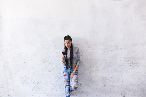 Young asian woman leaning back on blank wall and looking at smart phone. Wears ripped jeans and gray blouse, with long black hair.