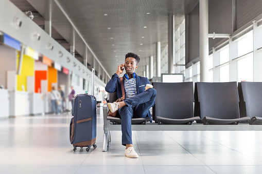 Young african businessman waiting for flight at the airport lounge and talking on mobile phone. Businessman in casuals sitting on chair and waiting for plane.