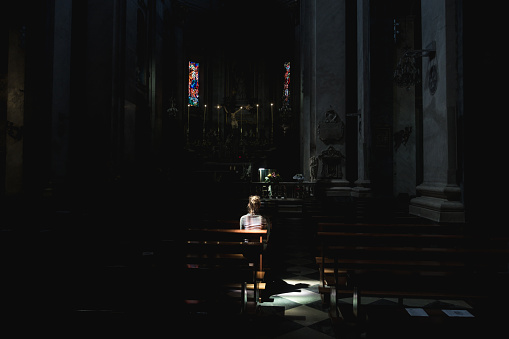 Young woman sitting and praying in a old Paroisse Elise Saint Jean Baptiste church in Bastia, Corsica. Beautiful light beams shining through the window illuminates her body.