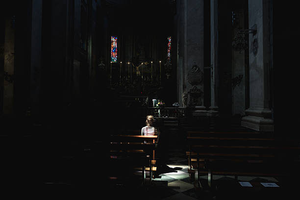 Young woman in church Young woman sitting and praying in a old Paroisse Elise Saint Jean Baptiste church in Bastia, Corsica. Beautiful light beams shining through the window illuminates her body. woman alone dark shadow stock pictures, royalty-free photos & images