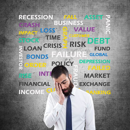 Frustrated young businessman wearing formalwear standing in front of wall with words of Recession, Crisis, Market, Stock, Risk, Loss, Income, Problem etc.