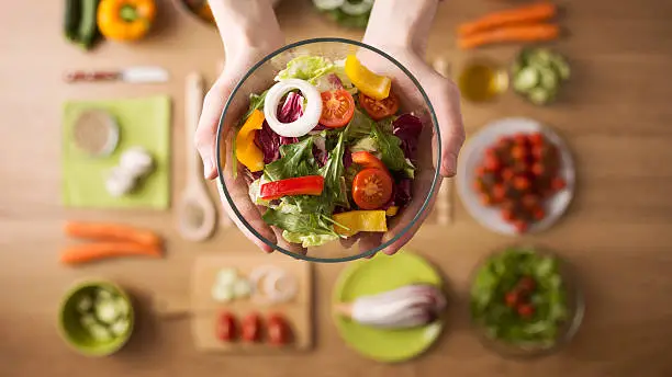 Hands holding an healthy fresh vegetarian salad in a bowl, fresh raw vegetables on background, top view
