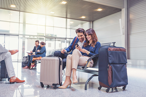 Young businessman and businesswoman sitting on bench at airport lounge using mobile phone, with people in background. People waiting for flight.