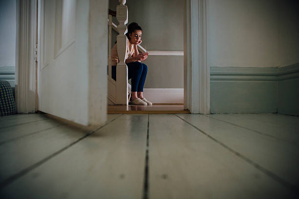Why is he not texting me back? Teenage girl sitting at the bottom of a staircase using a smartphone. sad 15 years old girl stock pictures, royalty-free photos & images
