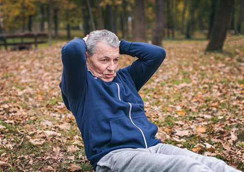 The photo of a senior man exercising. He wears blue sweatshirt and grey sweatpants. He is in shape of his life.