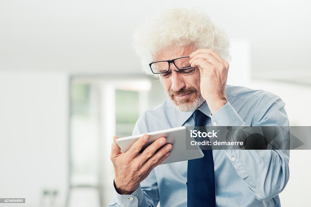 Businessman having eyesight problems Businessman having eyesight problems, he is using a tablet and adjusting his glasses, office interior on background Reading Stock Photo