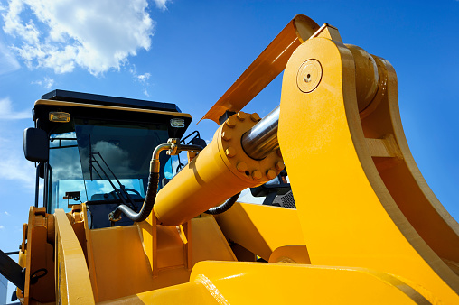 Bulldozer, huge yellow powerful construction machine with big scoop, focused on hydraulic piston arm, heavy industry, blue sky and white clouds on background