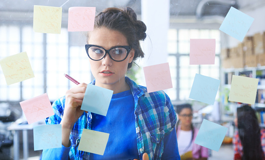 Young woman with eyeglasses writing on post-it notes on glass wall, in front of two female coworkers. All wear casual clothes.