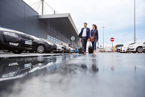 Full length outdoor shot of young man and woman pulling luggage. Businesspeople in an airport car parking.