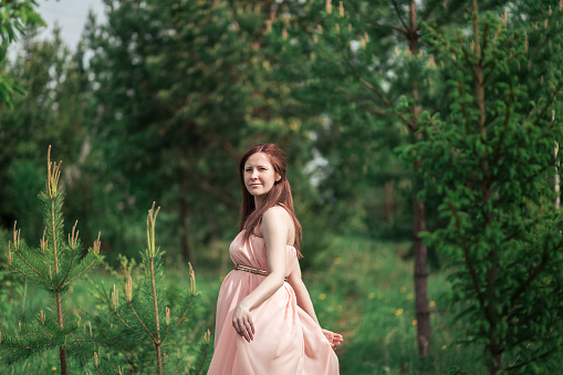 Pregnant young lady in a light pink dress in the woods in the summerYoung pregnant woman in pink dress in the woods in the summer