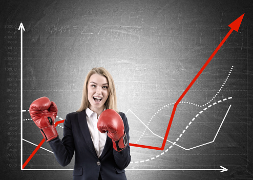 Cheerful woman in red boxing gloves is standing against blackboard with growing graphs on it. Concept of competition. Mock up