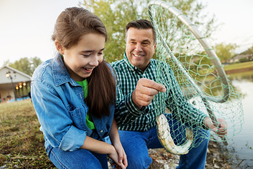 Cheerful grandfather smiles while holding a fish in a fishing net. His young granddaughter smiles proudly at the catch. The are sitting on the shore of a fishing pond.