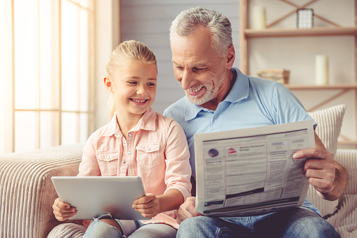 Cute little girl and her handsome grandpa are smiling while sitting on couch at home. Girl is using a tablet while grandpa is reading a newspaper