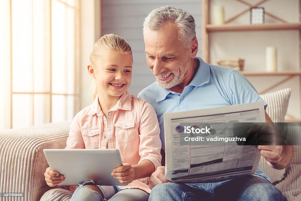 Grandpa and little girl at home Cute little girl and her handsome grandpa are smiling while sitting on couch at home. Girl is using a tablet while grandpa is reading a newspaper Newspaper Stock Photo