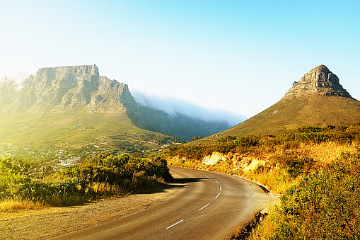 A winding road runs beside Lions Head and towards Cape Town's Table Mountain on a clear sunny day. The \
