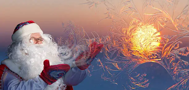 Santa Claus is blowing on window glass with frost and create amazing patterns, where the setting sun is reflected.