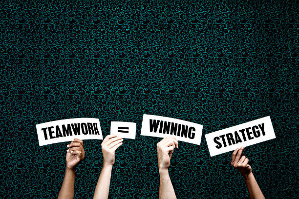 Four hands hold up words saying 'Teamwork = Winning Strategy' against a dark-patterned background.  Lots of copy space on the background above the words for your message.