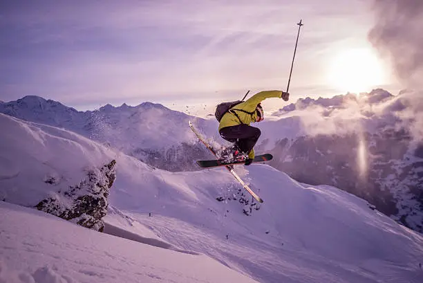 A skiier flies through the air on the ski slopes above Verbier in the Swiss alps. The mountains are blanketed in fresh snow and the sun is setting behind Mont Blanc and the Chamonix range.