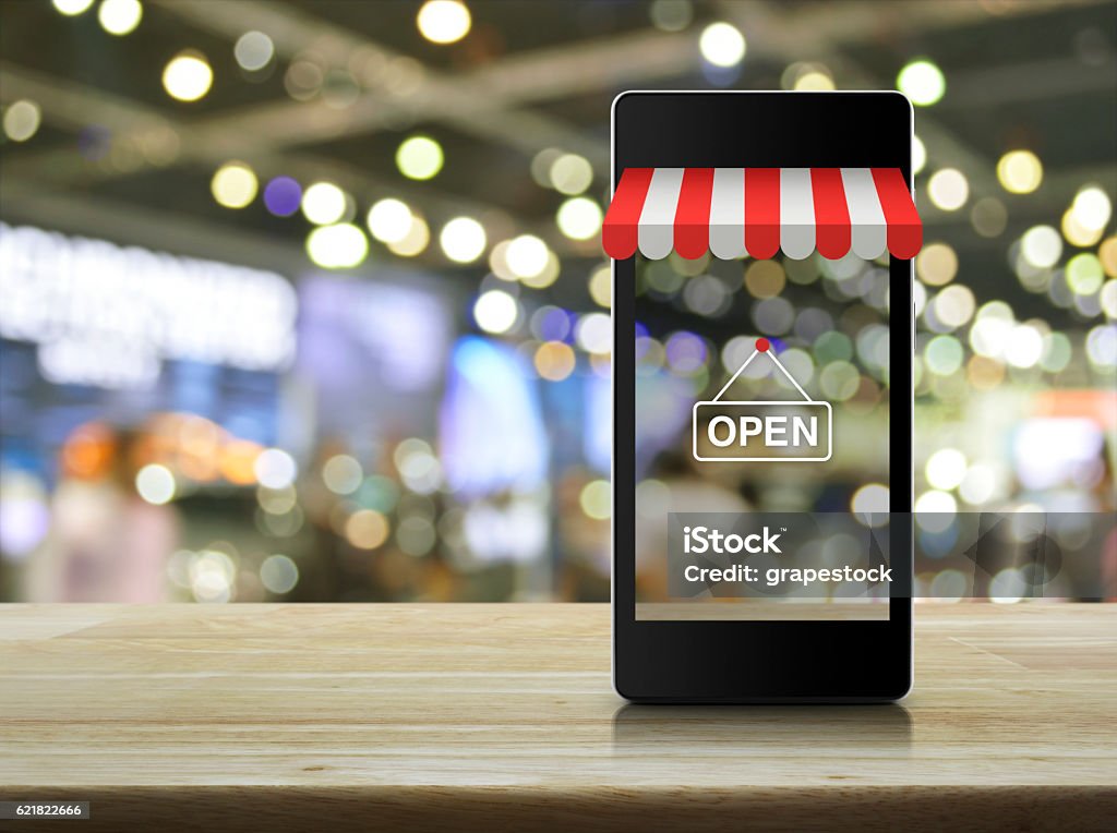 Modern smart mobile phone with on line shopping store graphic Modern smart mobile phone with on line shopping store graphic and open sign on wooden table over blur light and shadow of mall E-commerce Stock Photo