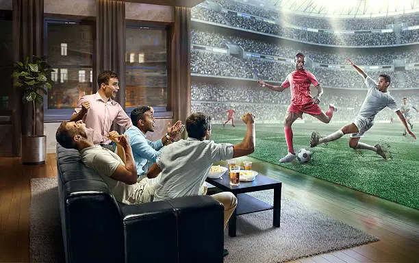 :biggrin:A group of young male friends are shocked while watching extremely realistic Soccer game on TV. They are sitting on a sofa in the modern living room faced to a real stadium with players instead of the front wall. It is evening outside the window.