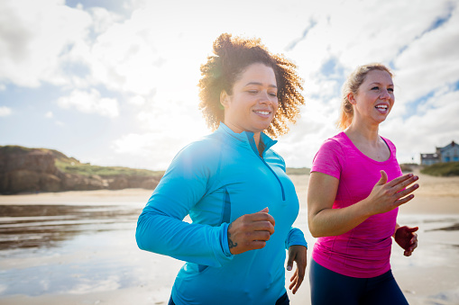 Two female friends running a long a beach together. They are jogging along the shoreline side by side and smiling