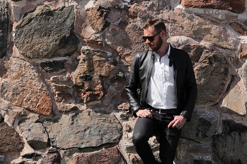 Attractive man wearing a black leather jacket and jeans with sunglasses, leaning back against a wall of stone outdoors on a sunny summer day.