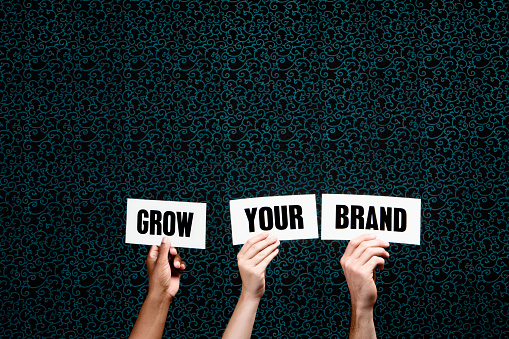 Three hands hold up words saying 'Grow Your Brand' against a dark-patterned background. Ample copy space on the dark background.