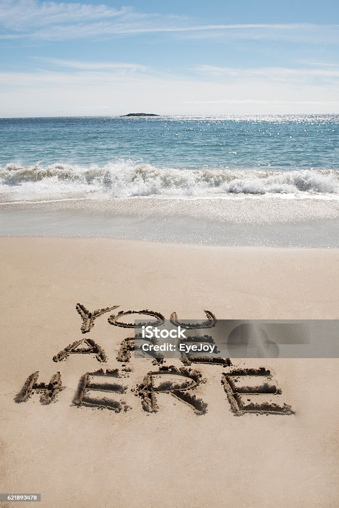 You are Here at the Beach These words, YOU ARE HERE, are written in the sand at a beach; waves, water and blue sky in background. Writing in sand entices others to come visit on a travel vacation. You Are Here Stock Photo