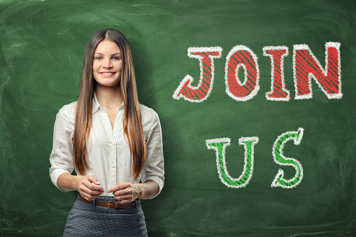 A young beautiful woman standing near the big bright words 'join us' written on the green chalkboard. Looking for the new members. We need you. The company is recruiting. An increase in the number of the staff .