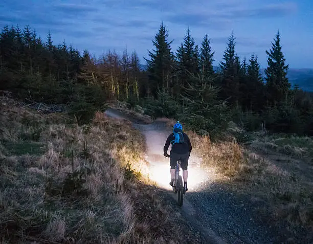 Rear view of a man using bright lights to illuminate the mountain bike trail in front of him as daylight fades.