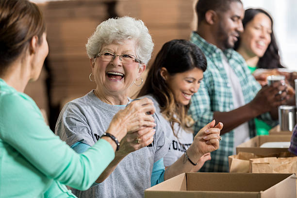 Beautiful cheerful senior woman volunteers at food bank Smiling Caucasian senior woman smiles while accepting a canned food donation at a charity food drive. Volunteers are working in the background. volunteer stock pictures, royalty-free photos & images