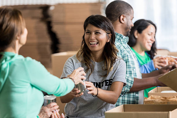 Volunteer accepts canned food donation at food drive Cheerful mid adult Hispanic female volunteer accepts a canned food donation during community food drive. She is wearing a gray volunteer t-shirt. Her friends are volunteering in the background. community stock pictures, royalty-free photos & images