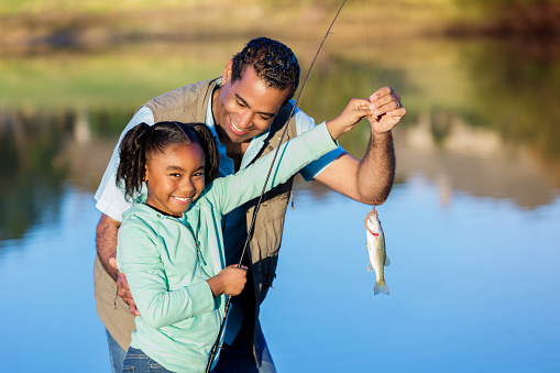 Pretty African American girls smiles while showing off a fish she caught with her grandpa. They are standing on the shore of a lake or pond.