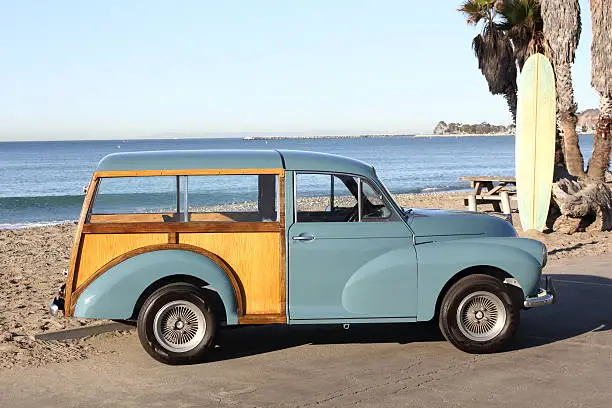 A Morris Minor Traveller at the beach with a surfboard in the background.