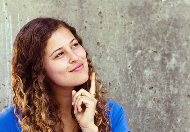 This beautiful young honey-blonde woman taps her chin as she thinks, smiling gently as she stands next to a grungy grey wall with copy space.