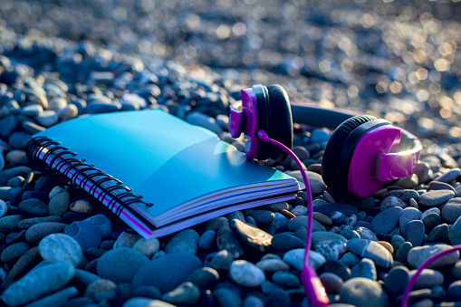 Pink headphones lying at paper notebook on sea beach. Closeup. Work and lifestyle concept. Freelancer. Musiic listening, Travel.