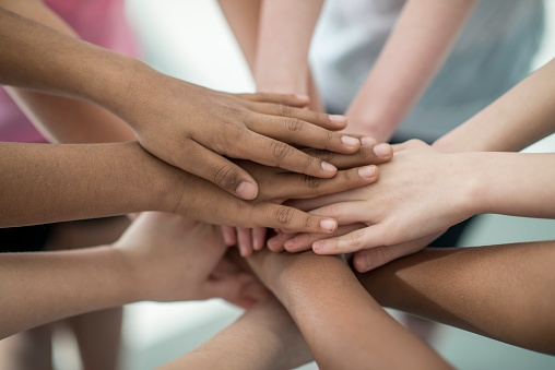 A multi-ethnic group of elementary age students are together in a huddle. The picture is of their hands stacked in the middle.