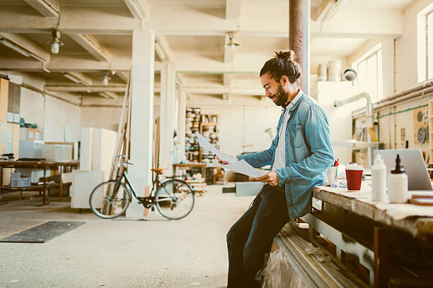 Young Male Carpenter Examining Blueprints In His Workshop Male carpenter standing in his workshop. He is making break and examining furniture drawings. Wearing headphones around neck. bike workshop stock pictures, royalty-free photos & images