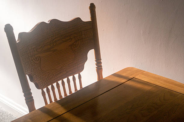 A simple chair and table bathed in natural light. Brown chair. Brown table. Natural lighting. White wall. Simple. Uncluttered.