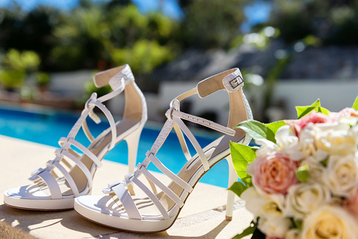 Horizontal color image of bridal white high heels sandal & bouquet of roses arranged next to the swimming pool.