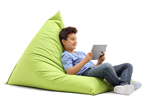 Joyful little boy sitting on a beanbag and looking at a tablet isolated on white background