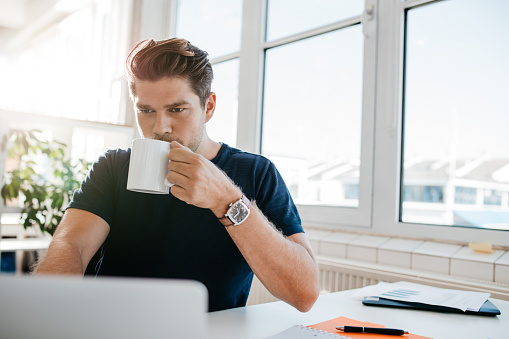 Young businessman drinking coffee and working on laptop at office. Male executive working at his desk and drinking coffee.