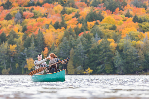 Happy couple canoeing in a lake in Canada. There are many trees on background with colourful leaves during autumn. They are young and happy, enjoying a canoe trip together. Wanderlust and nature concepts.