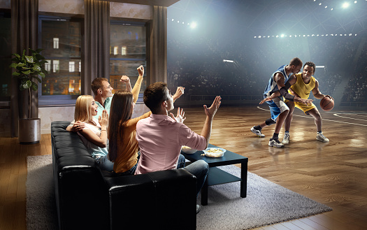 :biggrin:A group of young friends are shocked while watching extremely realistic Basketball game at home. They are sitting on a sofa in the modern living room faced to a real stadium with players instead of the front wall. It is evening outside the window.