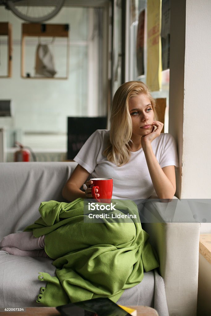 Woman resting in a couch. Girl holding a cup of coffee in hand and sad looking out the window. Adult Stock Photo