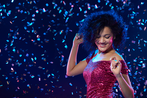 Happy mixed race woman in sequined dress dancing on a party over colorful background with confetti