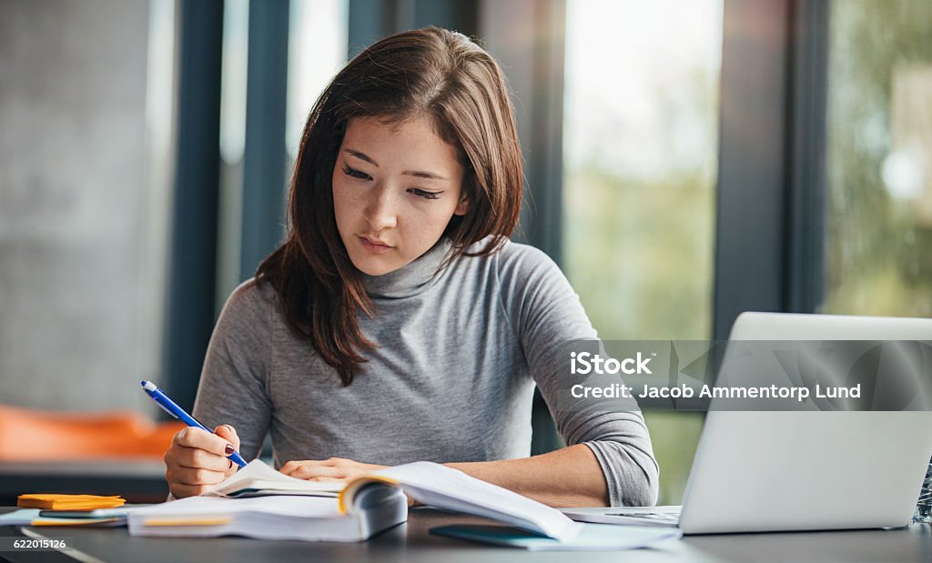 Woman taking down notes in diary Shot of young woman taking down notes in diary. Female university student preparing note for the exam at library. Educational Exam Stock Photo